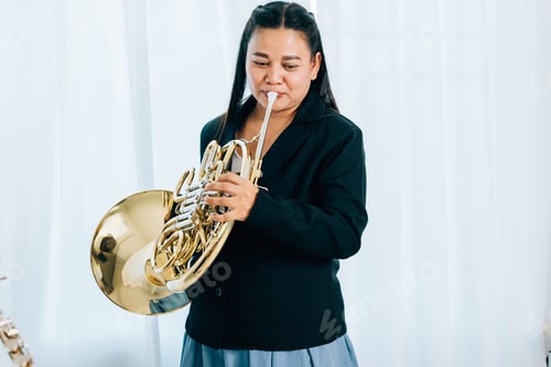 Preview: A woman stands at home in front of elegant curtains, playing a shiny gold horn. The warm lighting
