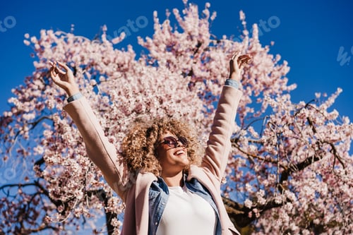 Preview: portrait of happy hispanic woman with afro hair in spring among pink blossom flowers. sunny nature