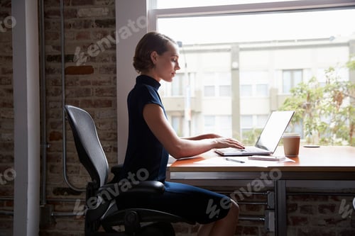 Preview: Businesswoman Sitting At Desk By Window Working On Laptop