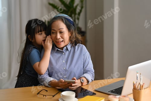 Preview: Adorable little girl whispering something to happy senior grandmother, spending leisure weekend.