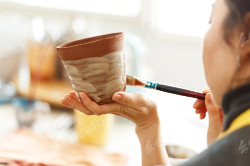 Preview: woman painting clay jug with colorfu paint shaping and sculpturing earthenware cup in pottery studio