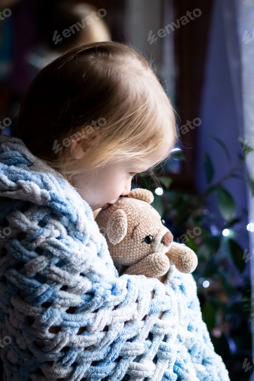 Preview: a little girl covered with a blanket gently kisses a teddy bear