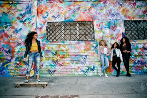 Preview: Four young women with curly hair standing in front of shutter covered in colourful graffiti, one