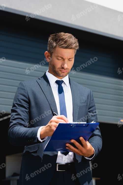 Preview: businessman in suit writing on clipboard with warehouse on background