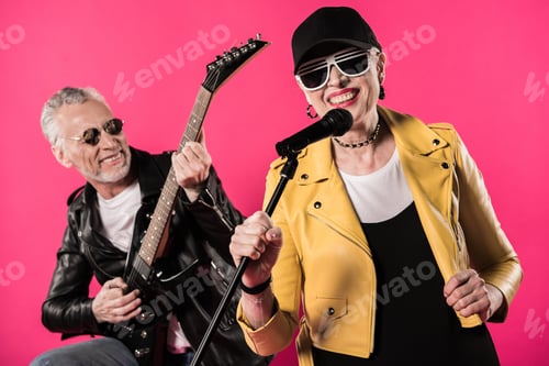 Preview: Smiling stylish senior couple performing rock and roll music isolated on pink