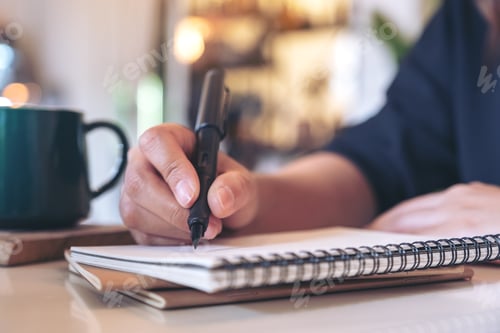 Preview: Closeup image of a woman's hand writing on blank notebook with coffee cup on table in cafe