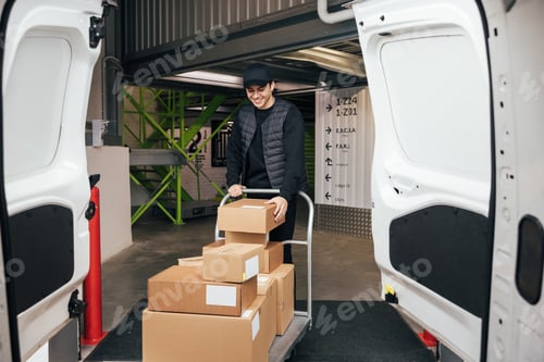 Preview: Smiling courier in uniform checking cardboard boxes on a cart while standing in a warehouse