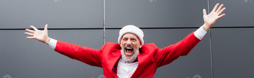 Preview: excited mature man in santa hat standing with outstretched hands near grey wall, banner