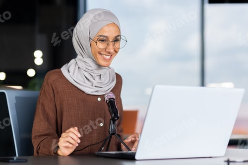 Preview: A young Muslim woman in a hijab is sitting in an office in front of a laptop and a microphone