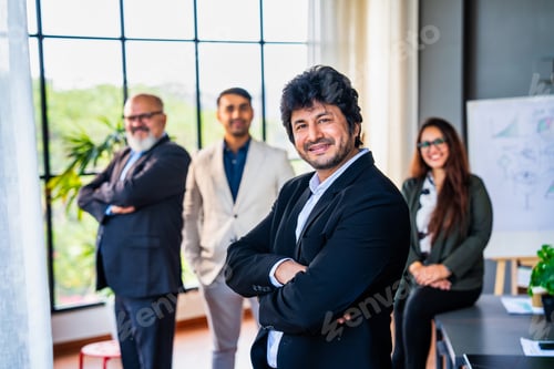 Preview: Confident Indian business leader standing with folded hands, colleagues out of focus in the office