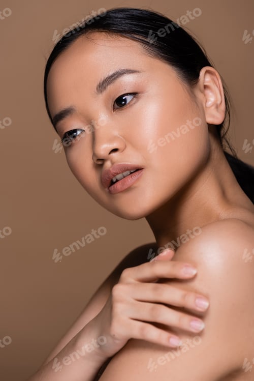 Preview: Young asian woman with natural makeup touching naked shoulder isolated on brown