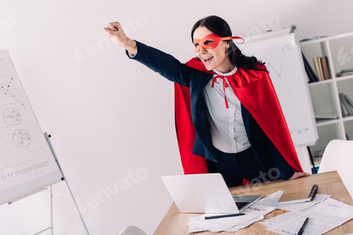 Preview: attractive super businesswoman in cape and mask screaming and standing with hand up in office