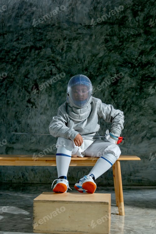 Preview: Vertical image of boy wear fencing sport uniform also hold epee and sit on wooden bench to relax.