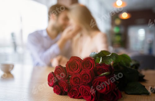 Preview: Bouquet of beautiful red roses on table in cafe and blurred young couple cuddling on background