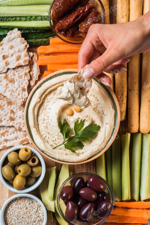Preview: partial view of woman and arranged hummus in bowl, pita bread, cut vegetables, dried tomatoes and