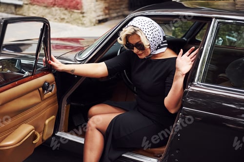 Preview: Blonde woman in sunglasses and in black dress going out from old vintage classic car
