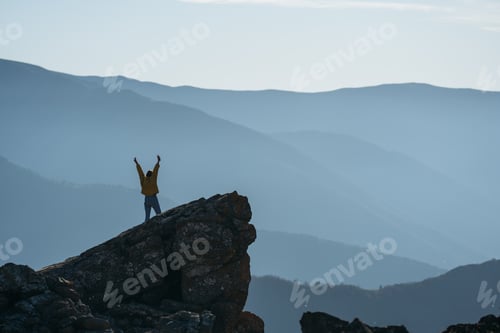 Preview: Happy woman in yellow jacket standing on rock with arms spread out. Space for text.