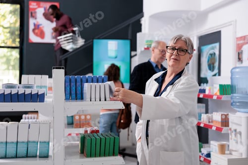 Preview: Senior drugstore worker looking at pills packages checking pharmacy shelves