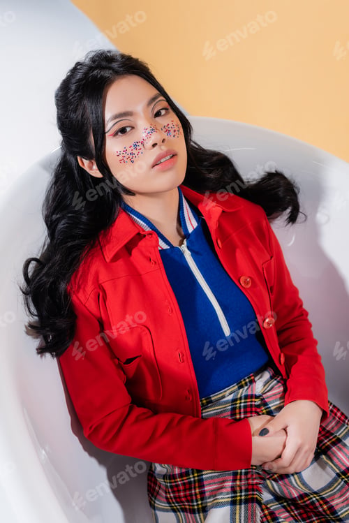 Preview: High angle view of trendy asian model looking at camera in bathtub on orange background