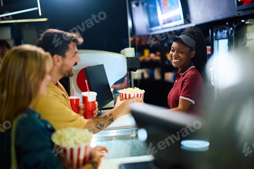 Preview: Happy black concession stand worker selling drinks and popcorn before movie projection in cinema.