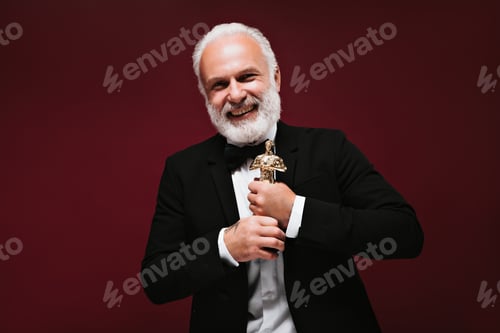 Preview: Cool man in black suit laughing and holding Oscar statuette. Smiling guy with grey beard and tattoo