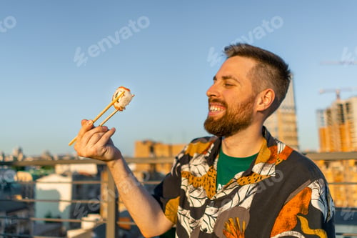 Preview: Close-up of a young attractive man with chopsticks in his hand eating sushi roll sitting on terrace