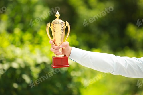 Preview: Young businessman holding gold trophy cup in green park, closeup