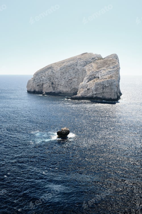 Preview: Vertical shot of a big rock in the middle of the sea with a blue sky in the background
