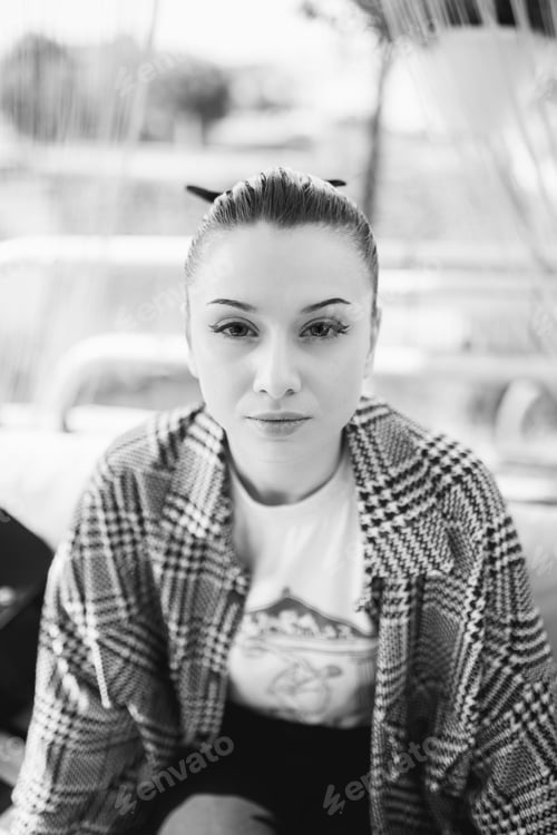 Preview: Vertical grayscale shot of a beautiful Caucasian girl wearing a checkered shirt, sitting outdoors