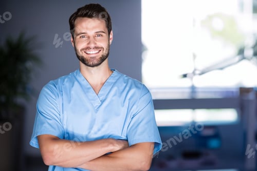 Preview: Man standing with arms crossed in hospital room, wearing light blue scrubs with plant and monitor