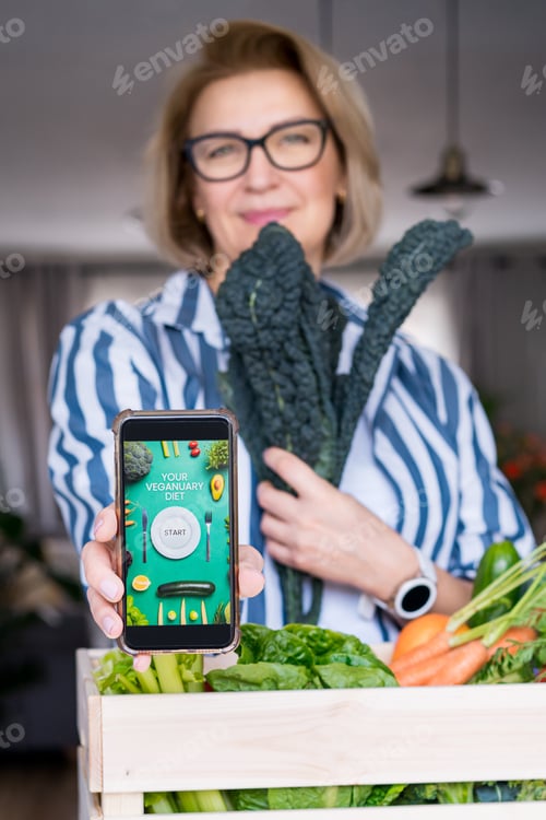 Preview: Woman holding phone with active online mobile app of Veganuary diet program and fresh kale