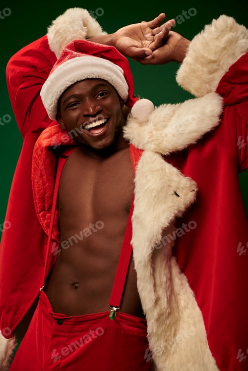 Preview: happy african american guy in christmas costume on shirtless body with hands behind head on green
