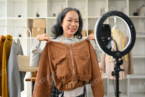 Preview: Senior female small business owner showing clothes and present detail during live streaming.