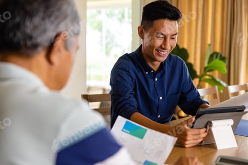 Preview: Happy diverse financial advisor and senior man discussing paperwork and using tablet in dining room