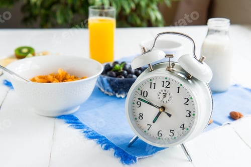Preview: Time for breakfast. Alarm clock with healthy food on the background. Bowl with cereals, nuts and ber