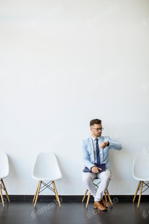 Preview: Young man sitting in the waiting room with a folder in hand and checking time before an interview