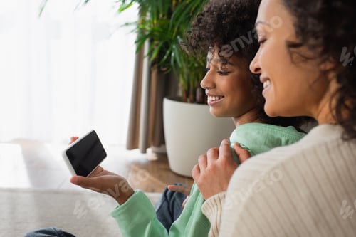 Preview: happy african american preteen girl holding smartphone with blank screen near positive mother