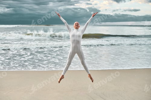 Preview: Joyful hairless girl with alopecia in white futuristic suit jumping sea sandy beach
