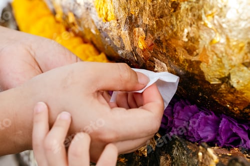 Preview: Close-up of a person's hands gently applying delicate gold leaf to a Buddha image or sacred object.