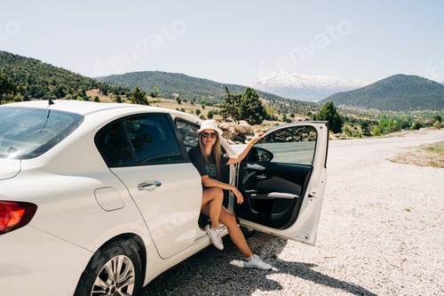 Preview: Woman traveler in hat sitting in car with mountain background. Road trip. Young adult woman in sun