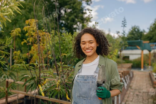Preview: Female gardener standing with hand trowel and looks camera on plants background