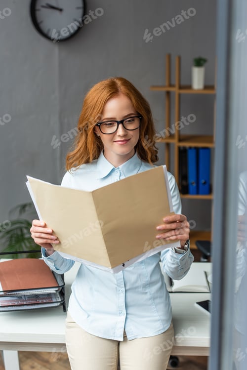 Preview: Smiling businesswoman looking at folder with paper sheets, while leaning on workplace with blurred