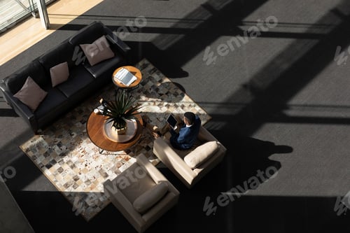 Preview: Man sitting on cream armchair wearing business casual and holding tablet in lounge, copy space