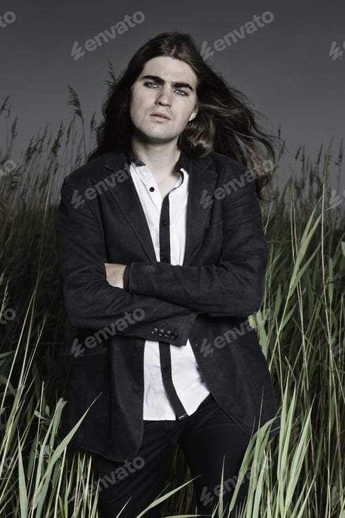 Preview: Young Man With Long Brown Hair Wearing Black Suit Standing In Field With Long Grass. Stormy Cloudy