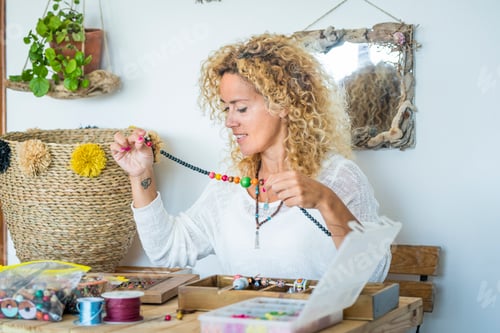 Preview: Happy woman at home work with beads and cords producing cheaper jewelry- modern female