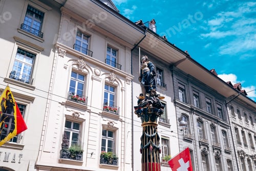 Preview: Historic fountain in Bern features intricate statues and vibrant flags under a clear sky