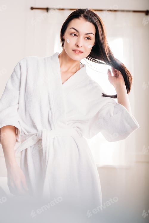 Preview: Young beautiful woman in white bathrobe with cream applied on her face, flipping her long hair