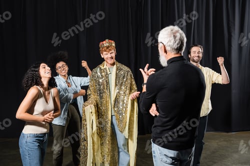 Preview: cheerful redhead man in king costume near multiethnic actors and art director on stage in theater