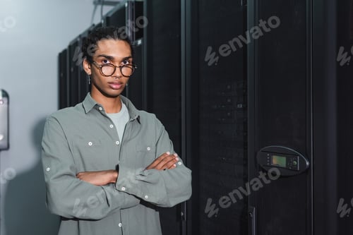Preview: african american programmer in eyeglasses standing with crossed arms and looking at camera in data