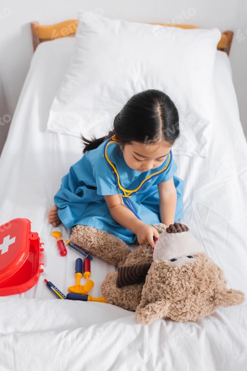 Preview: high angle view of asian girl examining teddy bear with toy stethoscope while playing on bed in